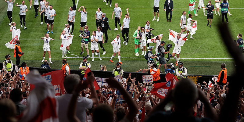 England players and staff celebrate on the Wembley pitch as fans hold up flags and banners, photo taken from the stands looking towards the pitch